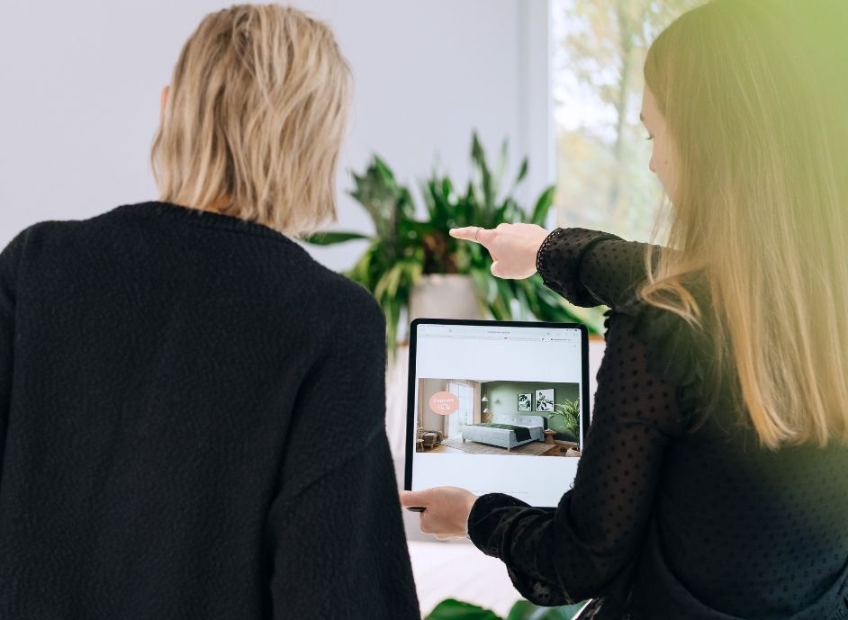 Zwei Frauen von hinten, eine zeigt auf einen Tabletbildschirm mit einem Schlafzimmerfoto