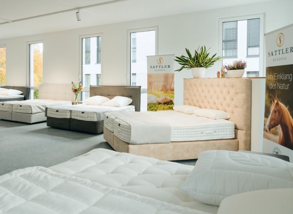 Mehrere Betten mit unterschiedlichen Kopfteilformen in einem hellen Ausstellungsraum mit großen Fenstern und Pflanzen